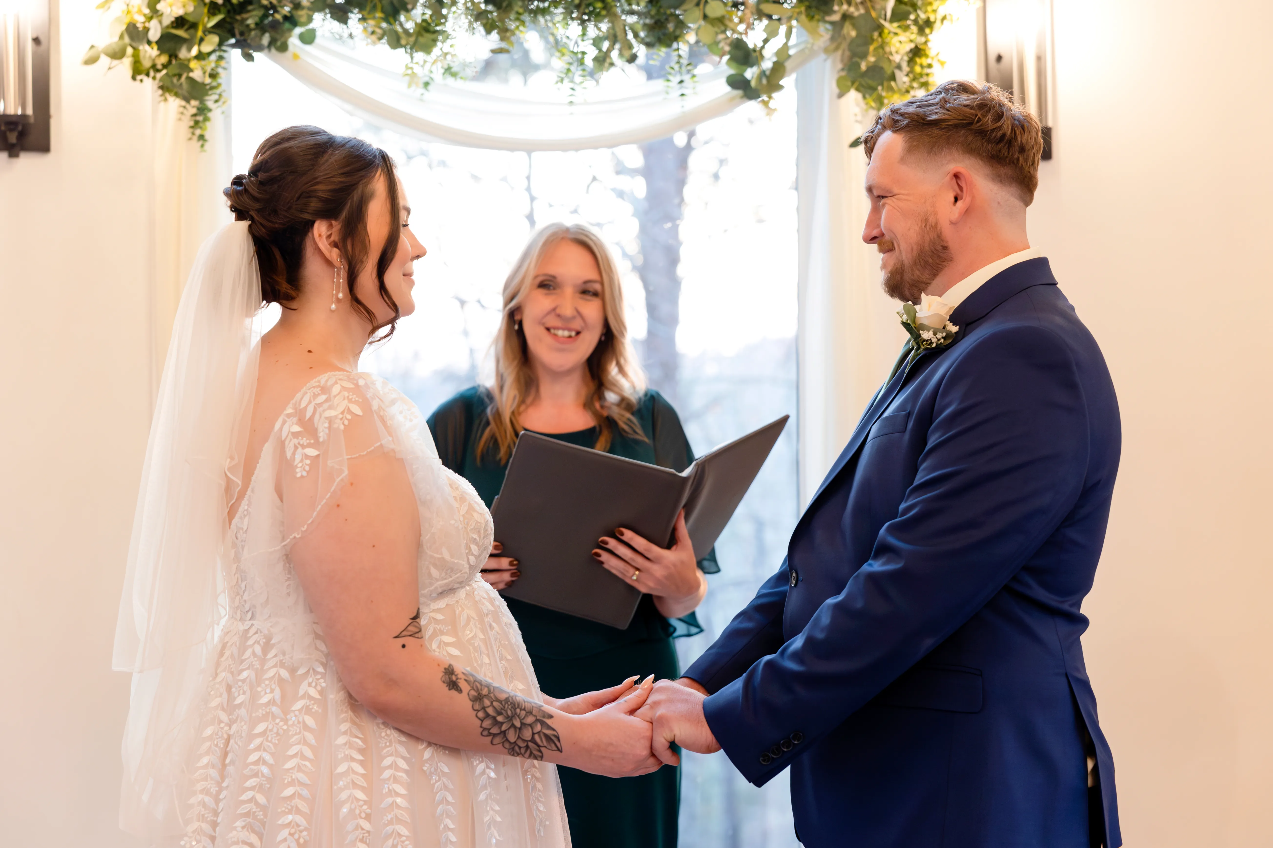 Bride and groom holding hands during ceremony vows under a floral arch